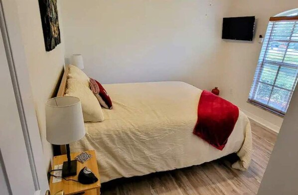 A bedroom with a bed, a bedside lamp, a TV on the wall, and a window with blinds.