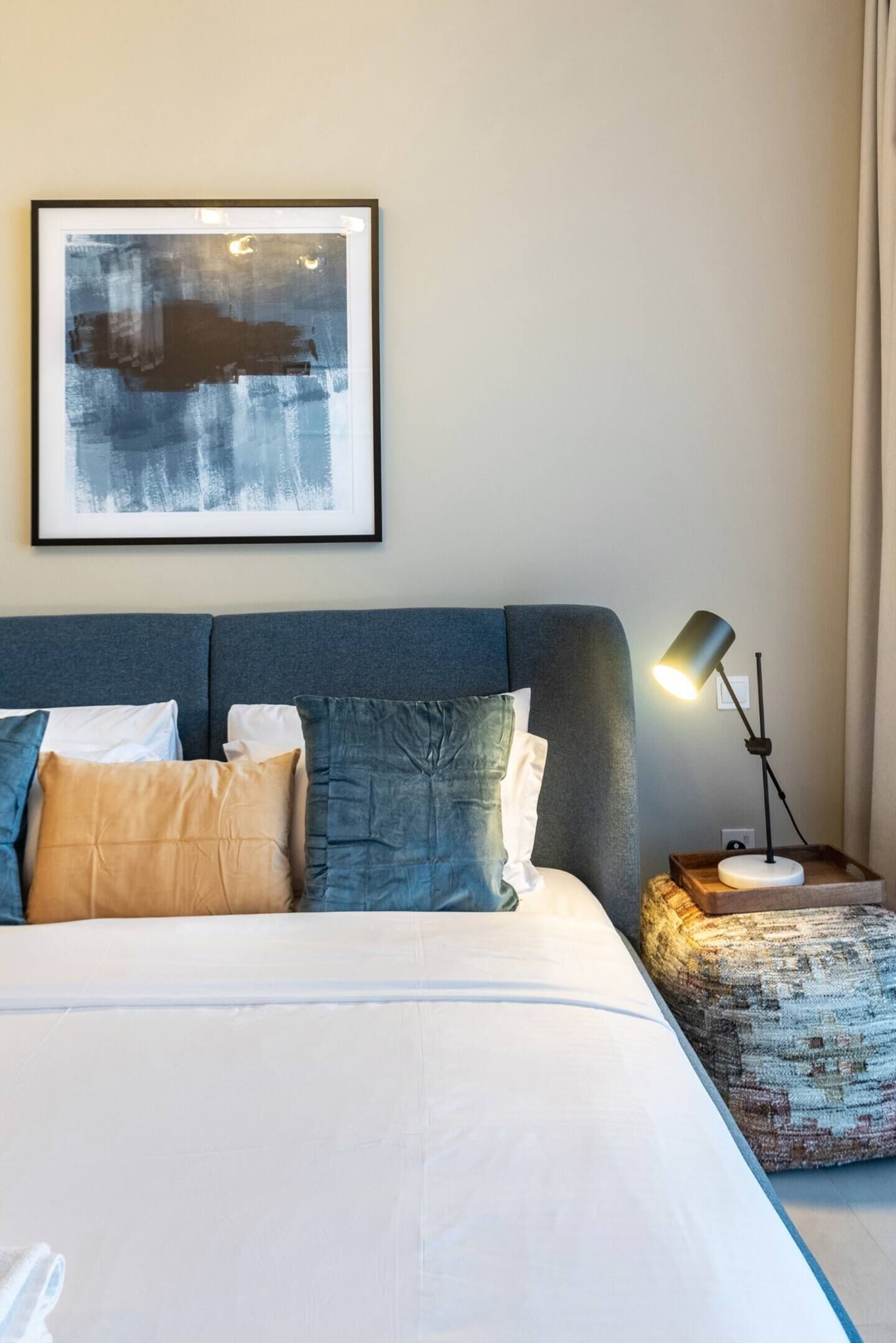 A neatly made bed with a white comforter and multiple pillows, a framed abstract painting on the wall, and a bedside table with a lamp.