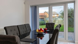 A dining area with a glass table and chairs, a bowl of fruit, and a view of a garden and house through large windows. - 4 Luxurious Executive Villa (Sheerness)