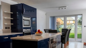 A modern kitchen with dark blue cabinets, a white countertop, and a bowl of fruit. There is a dining area with a table and chairs, and a sliding glass door leading to a garden. - 4 Luxurious Executive Villa (Sheerness)