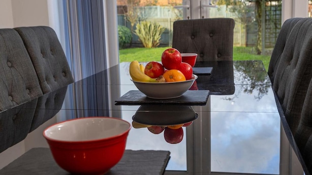 A dining area with a glass table, two grey upholstered chairs, a red bowl, and a bowl of fruit on the table. - 4 Luxurious Executive Villa (Sheerness)