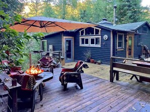 Una terraza de madera con mobiliario exterior, un hogar para fuego y una zona cubierta con una sombrilla.