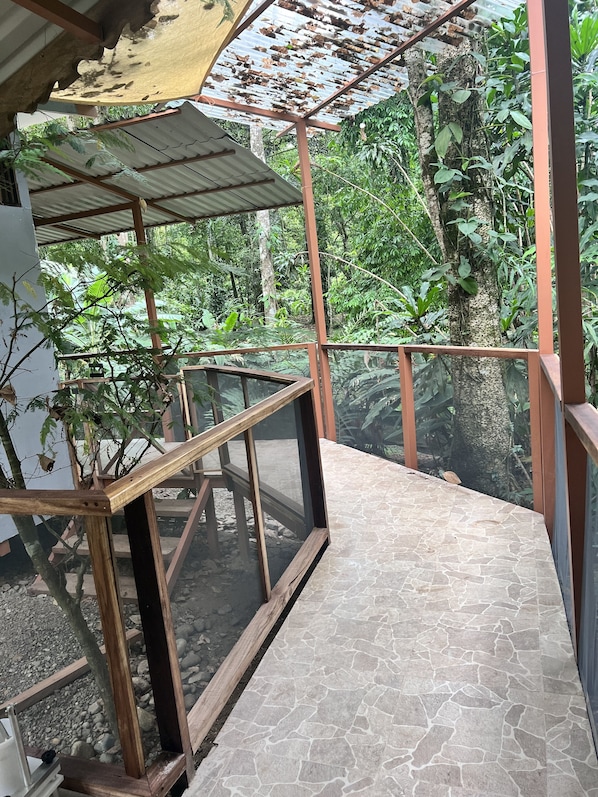A glass-enclosed balcony with a stone-tiled floor, surrounded by lush greenery and a metal roof structure.
