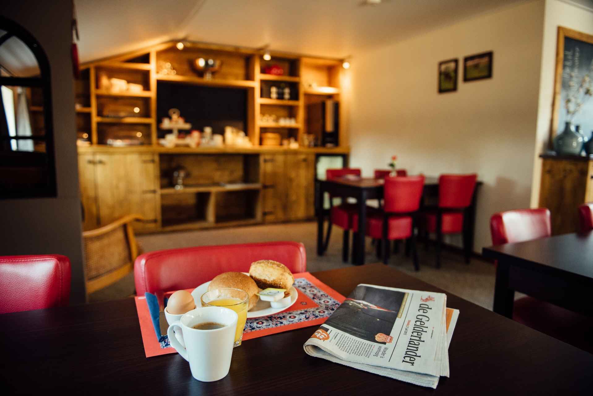 Uma sala de jantar com mesa posta para o café da manhã, incluindo ovos, um croissant e uma xícara de café, com um jornal sobre a mesa.