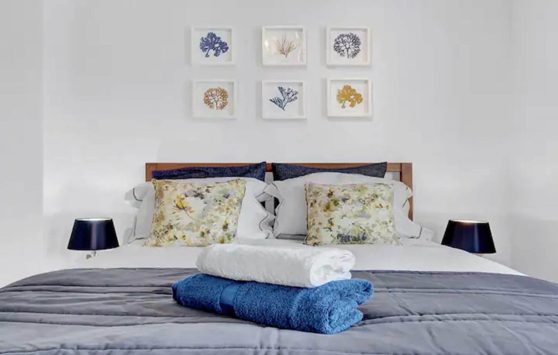 Un lit bien fait, agrémenté d’oreillers à motifs floraux, d’une couverture pliée aux couleurs blanc et bleu, et d’une couette grise. On y trouve deux lampes de chevet et six reproductions botaniques encadrées accrochées au mur.