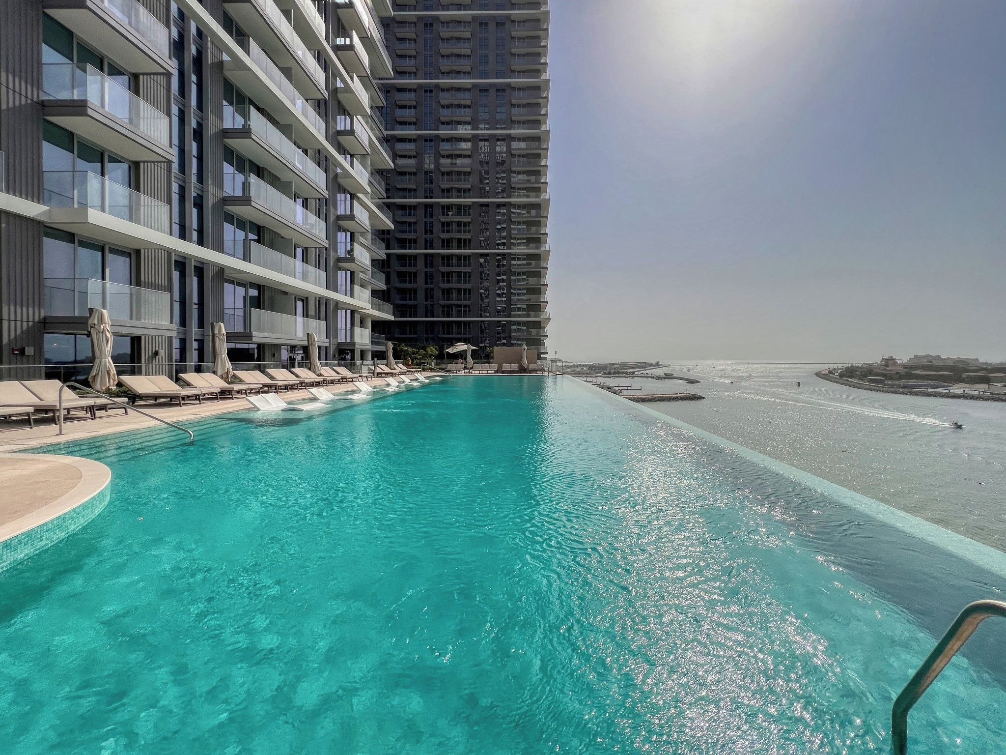 Ein Hotelpool mit klarem, blauem Wasser, umgeben von Liegestühlen und Sonnenschirmen, mit Blick auf das Meer und einem Hochhaus im Hintergrund.