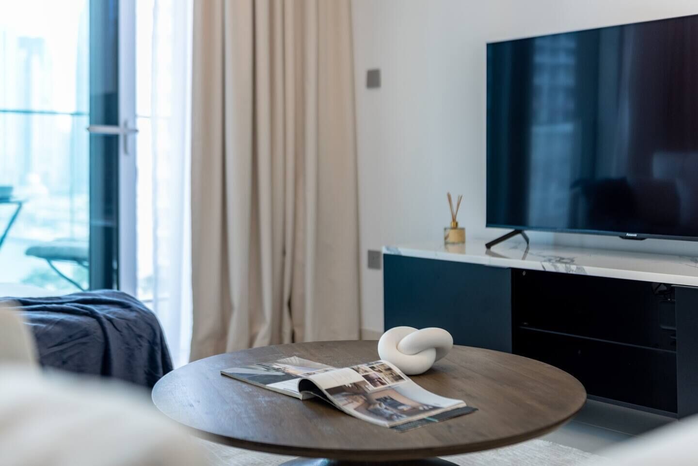 Ein modernes Hotelzimmer mit einem runden Holztisch, einem flachbildfernseher an der Wand und einem Fenster mit Vorhängen.
