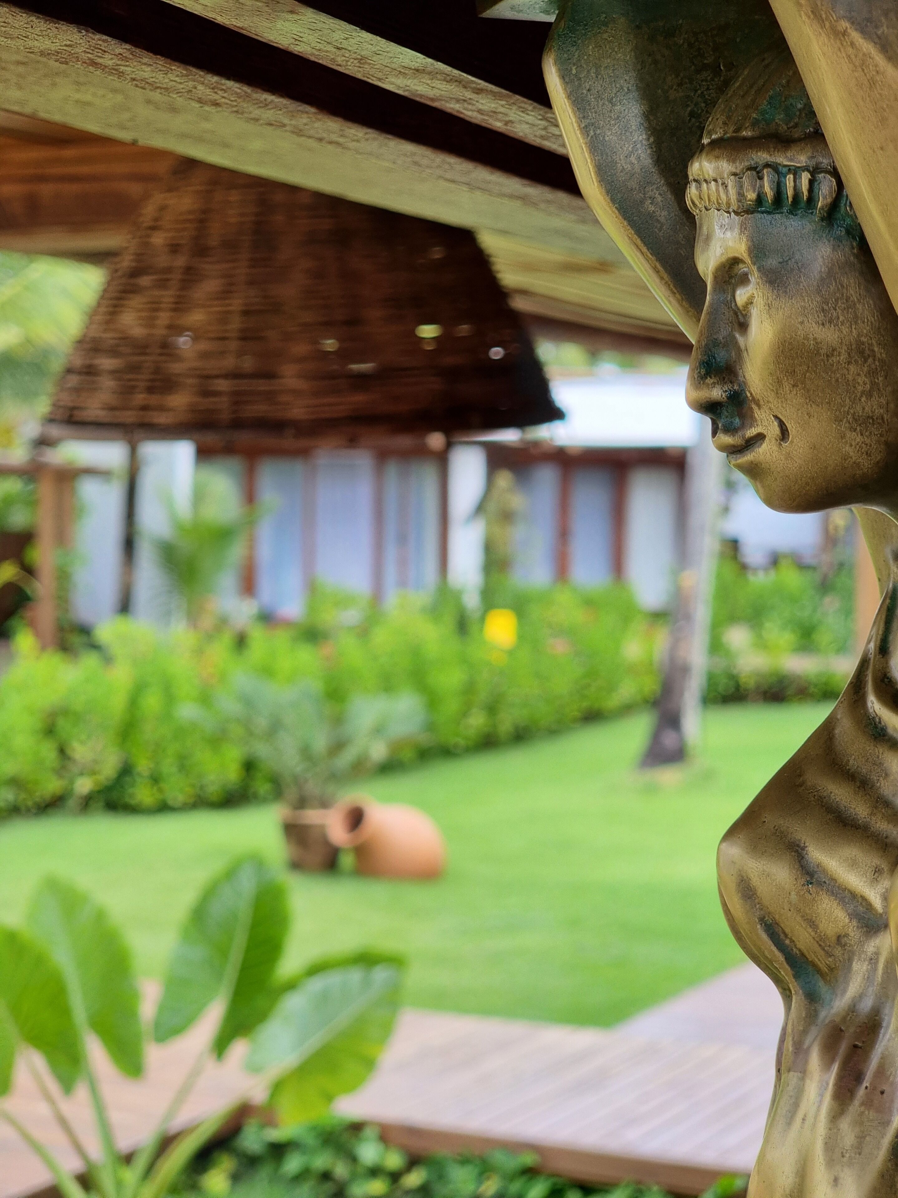 A garden with a statue, a thatched-roof structure, and a clay pot.