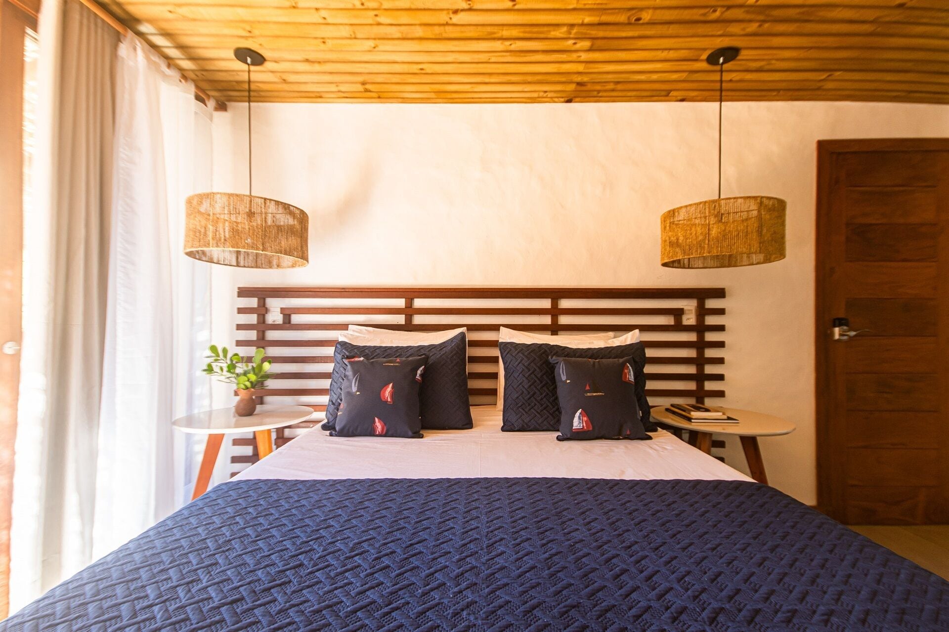 A bedroom with a wooden headboard, two hanging wicker lamps, a round side table with a plant, and a dark blue quilted bedspread.