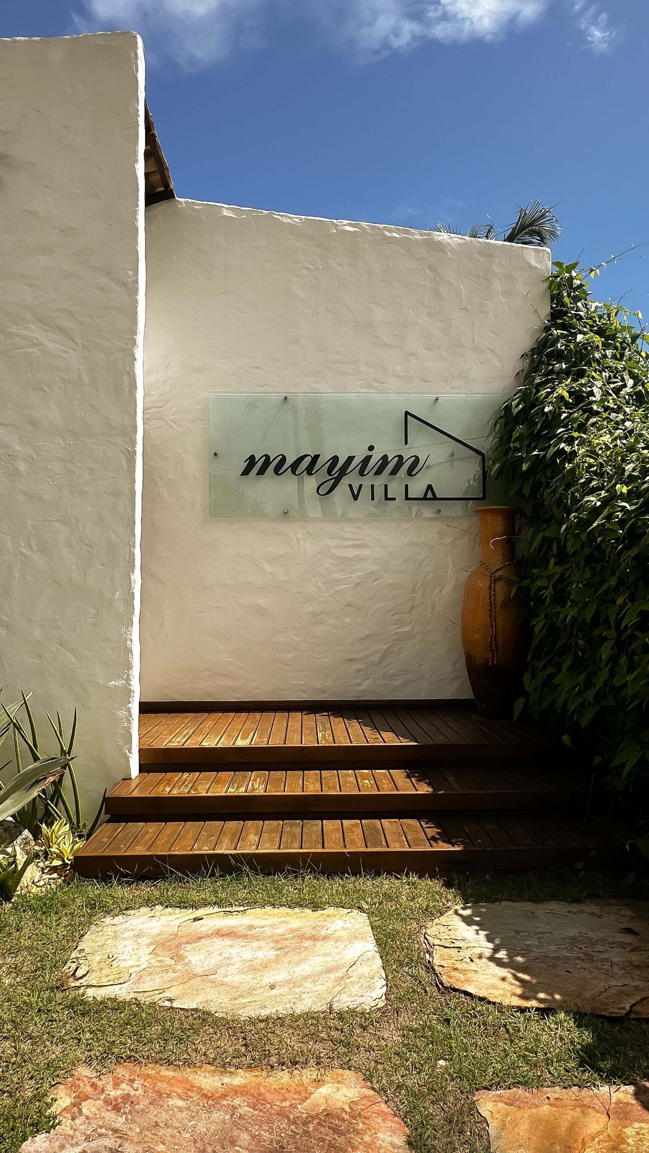 Entrada a una villa con un cartel que dice "mayim villa", escalones de madera y un jardín con césped y rocas.