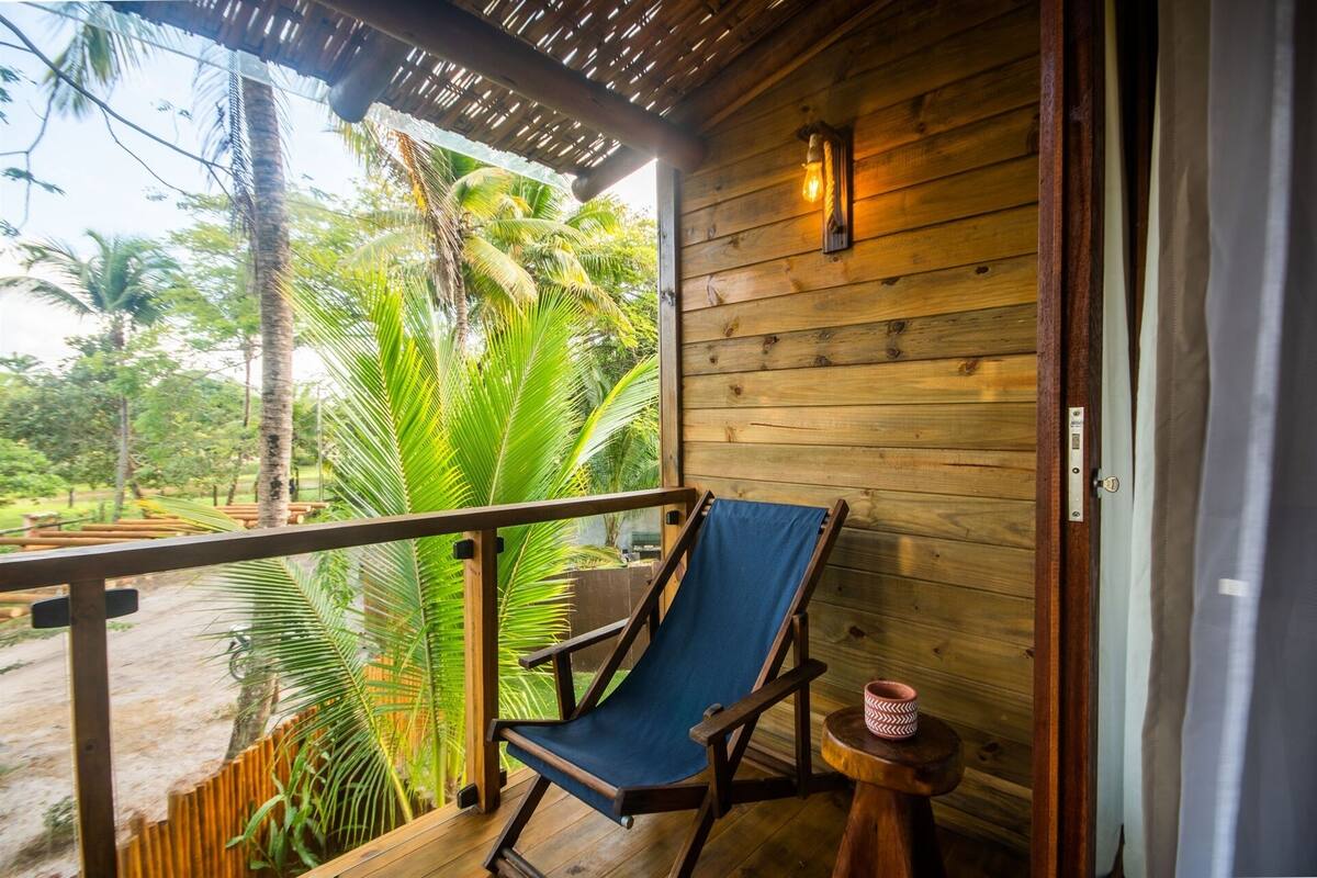A wooden balcony with a blue cushioned chair, a small round table with a patterned mug, and a view of lush green palm trees.