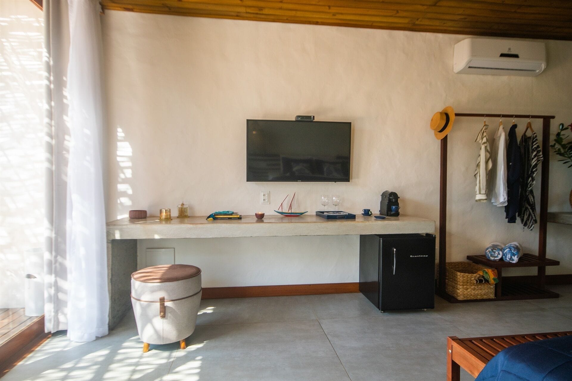 A compact living space with a TV mounted on the wall, a small kitchenette, a seating area with a bench, and a clothing rack.