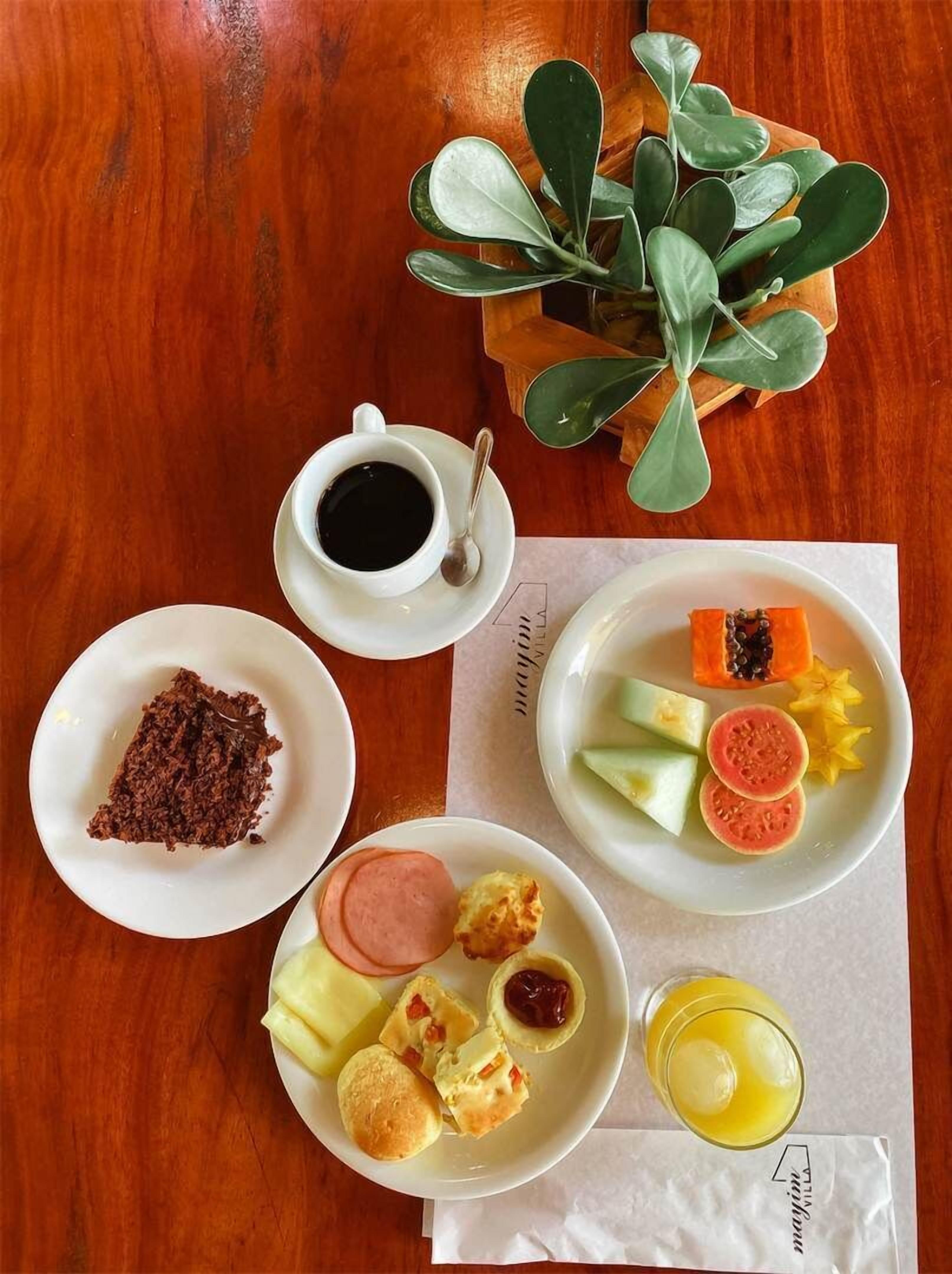 Une table en bois sur laquelle on trouve une variété d’aliments, notamment une tasse de café, une assiette de fruits coupés, une assiette de pâtisseries et un petit bol de chocolat.