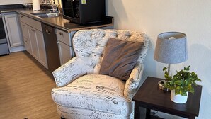 A living room with a patterned armchair, a small side table with a lamp and plant, and a kitchen area in the background. - The Asbury House Guest Rentals (Moscow)