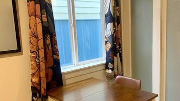 A dining area with a wooden table and two chairs, a window with curtains, and a door in the background.