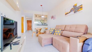 A modern living room with a large flat-screen TV, a beige sofa, and a dining area with chairs and a table. - GB67 Ótimo Apto 2 Suítes - Guarajuba (Camaçari)