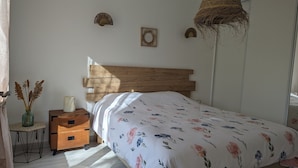 A bedroom with a wooden headboard, a floral bedspread, a bedside table with a vase of dried flowers, and a small wall-mounted shelf. - Maison à 50m de L'océan (Yves)