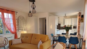 A living room with a yellow sofa, wicker chairs, and a dining table with blue chairs. There is a kitchen in the background with white cabinets and a wooden shelf with flowers. - Maison à 50m de L'océan (Yves)