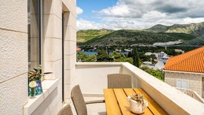 A balcony with a wooden table and wicker chairs, overlooking a mountainous landscape with a lake and buildings. - Villa Diana: Bayside View Beauty - a romantic home
 (Dubrovnik)