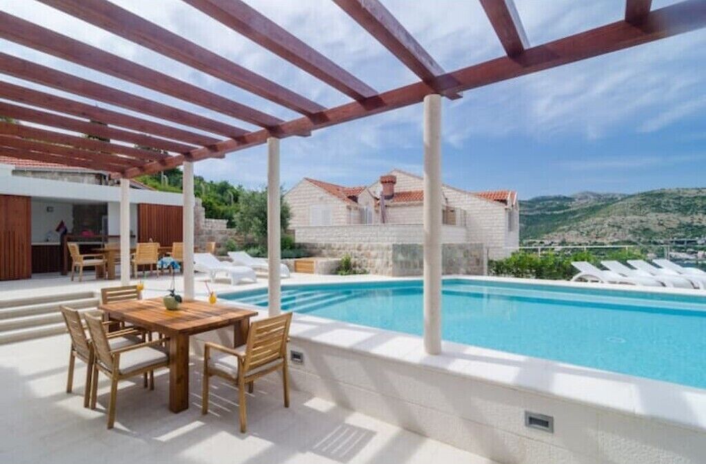 Uma área à beira da piscina com um pérgola de madeira, uma mesa de jantar com cadeiras e vista para um edifício no alto de uma colina.