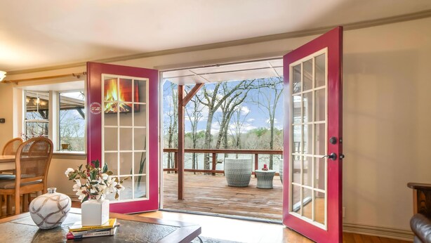Una habitación con terraza de madera, puerta roja y vistas a árboles y agua.