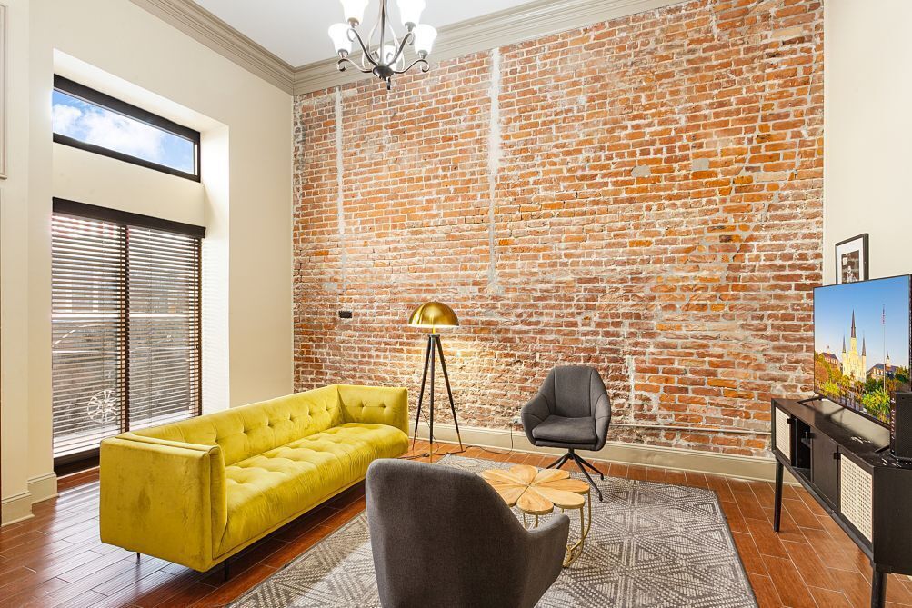 A living room with a brick wall, a yellow sofa, a grey armchair, a floor lamp, and a TV stand.