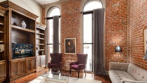 A living room with a brick wall, two armchairs, a sofa, a glass coffee table, and a bookshelf with a TV.