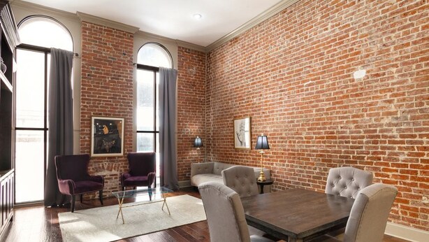 A room with a brick wall, a wooden dining table with four chairs, two armchairs, and a small side table.