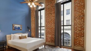 A bedroom with a large bed, a ceiling fan, a window with curtains, and a brick wall.