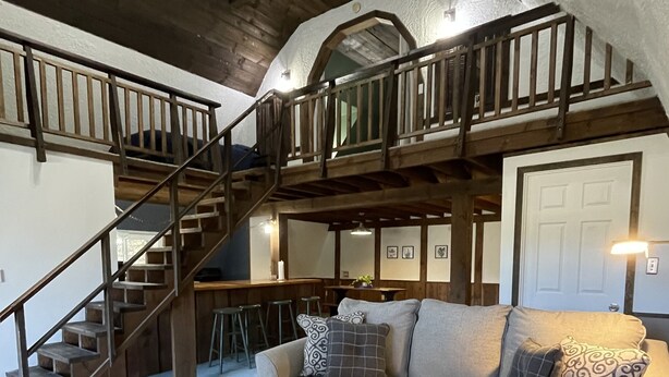 A spacious loft with a high ceiling, exposed wooden beams, a staircase, a cozy seating area with a sofa and a round coffee table, and a kitchen area in the background.