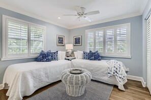 A bedroom with two beds, a ceiling fan, and large windows with shutters.