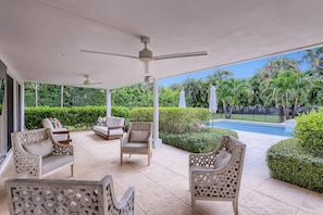 Area relax esterna coperta con sedie e ventilatore a soffitto, affacciata su una piscina e vegetazione lussureggiante.