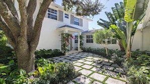 Una casa de dos pisos con un patio empedrado, rodeada de exuberante vegetación y un árbol grande.