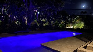 A rectangular swimming pool with a blue-lit interior, surrounded by a stone deck and lush vegetation. - Casa del Mundo 2bd 3.5ba With Private Pool (West Palm Beach)