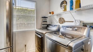 A compact laundry room with a stainless steel washer and dryer, a window with blinds, and a shelf with cleaning supplies. - Casa del Mundo 2bd 3.5ba With Private Pool (West Palm Beach)