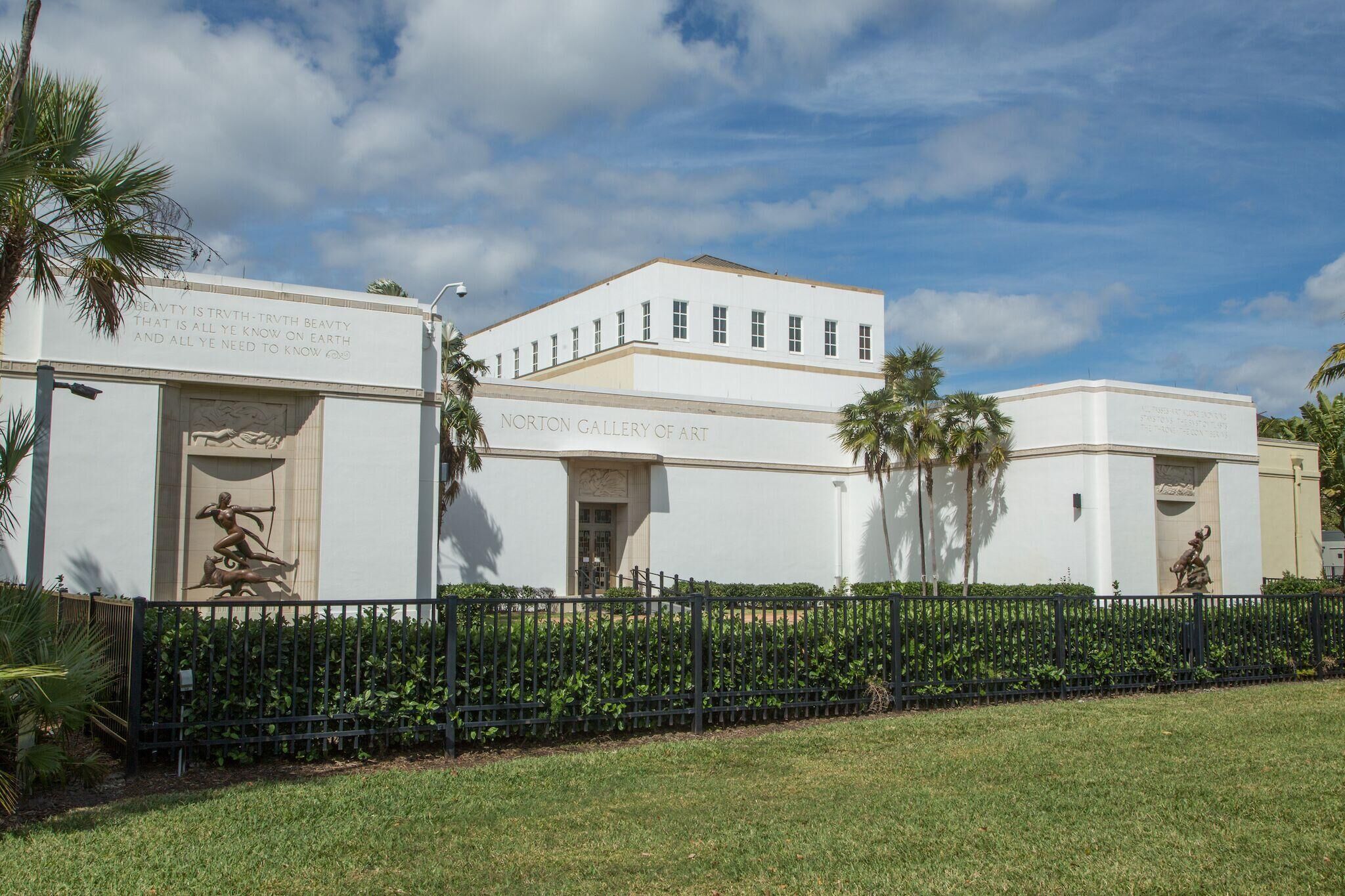 The exterior of the Norton Gallery of Art, featuring a white building with a flat roof, a black fence, and palm trees.
