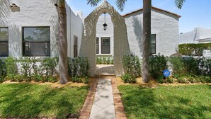 A white building with a stone archway entrance, a paved pathway, and a small garden with palm trees and shrubs. - Casa Sunshine NEW (West Palm Beach)