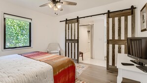 A bedroom with a bed, a desk, a TV, a ceiling fan, and a sliding door leading to a bathroom. - Cortez Bungalow, 3 Bd 2 Ba, Private Pool (West Palm Beach)