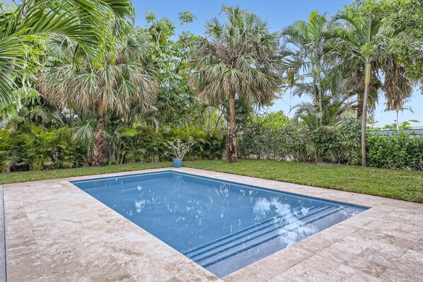 A rectangular swimming pool with a tiled edge, surrounded by lush greenery and palm trees.