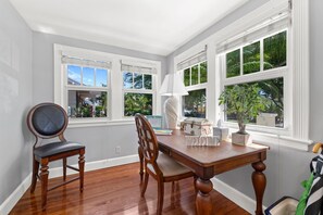 A dining area with a wooden table and chairs, large windows with blinds, and a view of greenery outside.