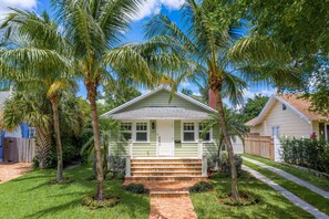 A single-story house with a front porch, a brick pathway leading to the entrance, and palm trees surrounding the property.