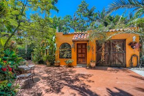 Quaint building with a rustic facade, wooden door, and arched window, surrounded by lush greenery and a shaded patio.