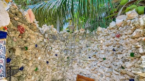 Zona de ducha con paredes de piedra y puerta de madera, rodeada de hojas de palmera y diversas conchas.