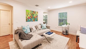 A living room with a sofa, a coffee table, a colorful abstract painting, and a window with blinds. - Key Lime Bungalow 2 BD 1 BA With Private Pool (West Palm Beach)