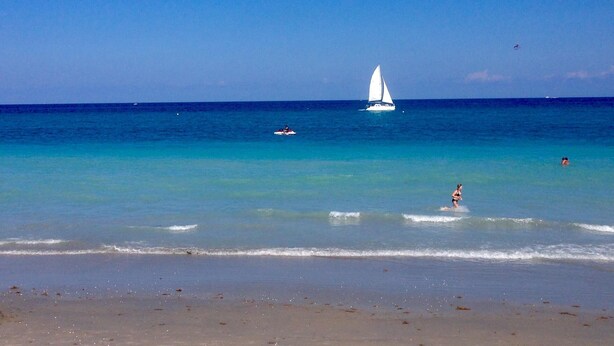 A beach with clear turquoise water, a sailboat, and a person surfing.
