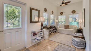 A spacious living room with a white door, a bookshelf, a desk, a white sofa, and a ceiling fan. - Nini's Cottage 4bd 3ba Vacation Rental (West Palm Beach)