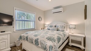 A bedroom with a bed, nightstands, a TV, a window with blinds, an air conditioner, and a wall-mounted clock. - Nini's Cottage 4bd 3ba Vacation Rental (West Palm Beach)