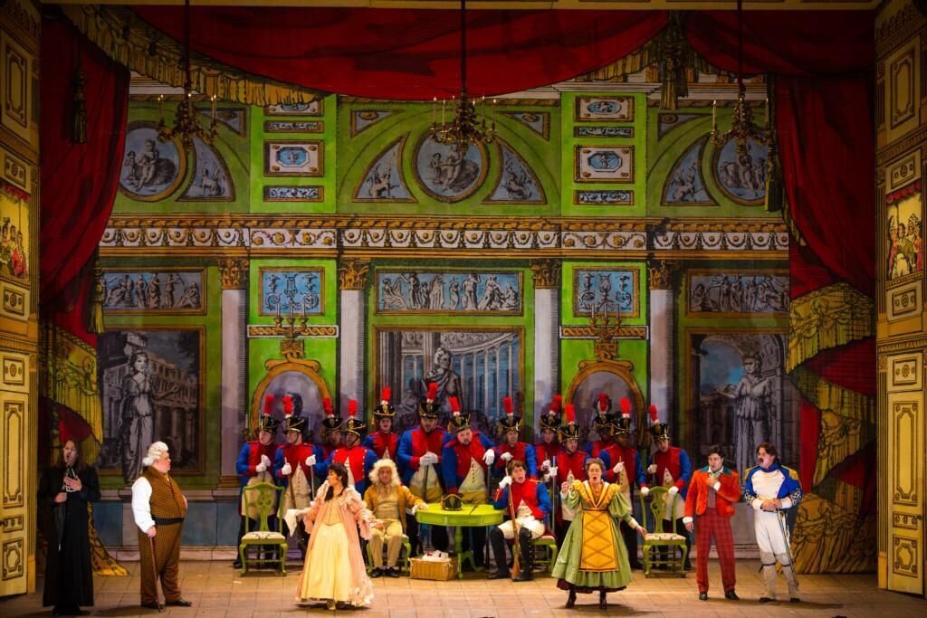A stage with a group of people in period costumes, a chandelier, and classical paintings on the walls.