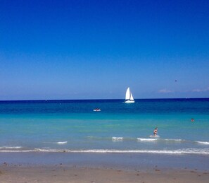 Una spiaggia con acqua turchese cristallina, una barca a vela e una persona che fa surf.