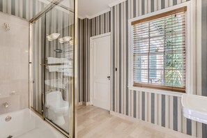 A bathroom with a glass shower enclosure, a white toilet, and a window with blinds.